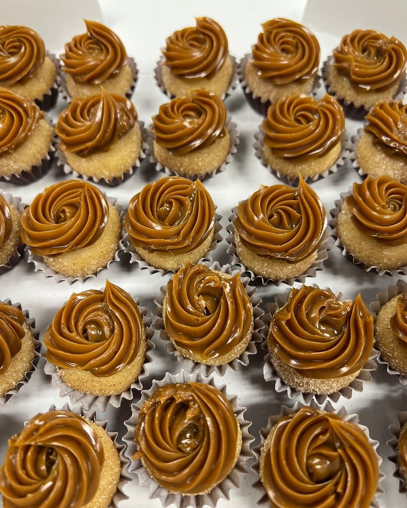 Rows of small, round desserts with swirled caramel frosting in fluted paper cups.