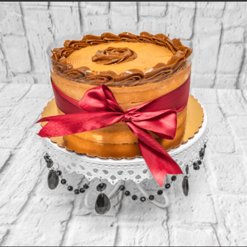 A caramel cake with a red ribbon on a white decorative stand against a white brick wall.