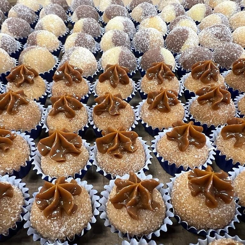 Rows of brigadeiros, some topped with dulce de leche, others coated in coconut or chocolate sprinkles.